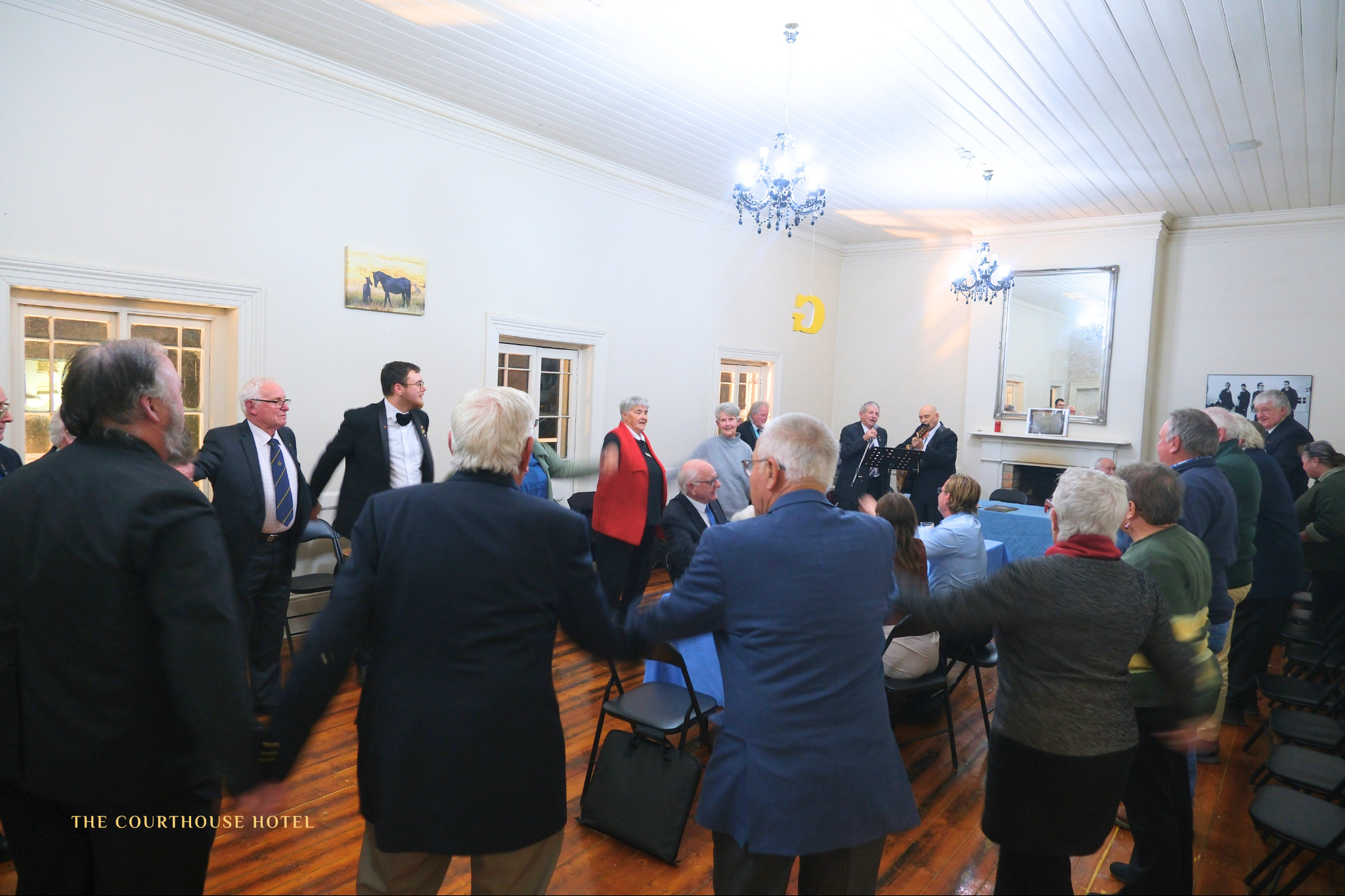 Guests standing in a circle enjoying live music in the upstairs function room at The Courthouse Hotel Smythesdale, with timber floors, high white ceilings and chandeliers.